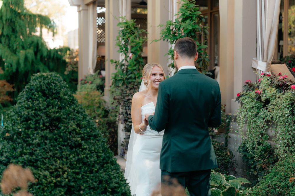 A romantic wedding ceremony takes place in a lush garden setting with elegant stone columns and greenery.