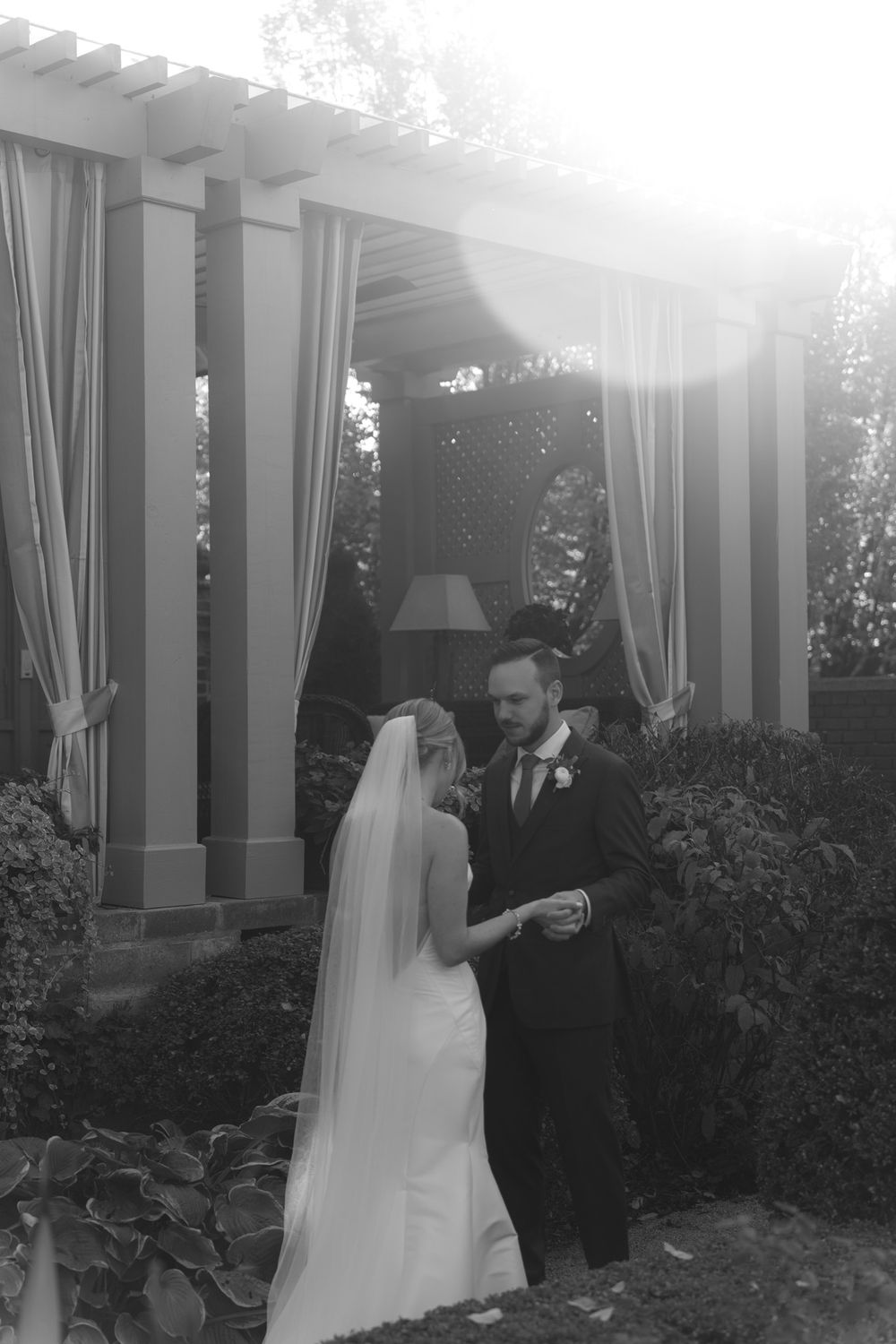 A black and white wedding photo showing a couple standing under an outdoor pergola with white curtains and garden surroundings.