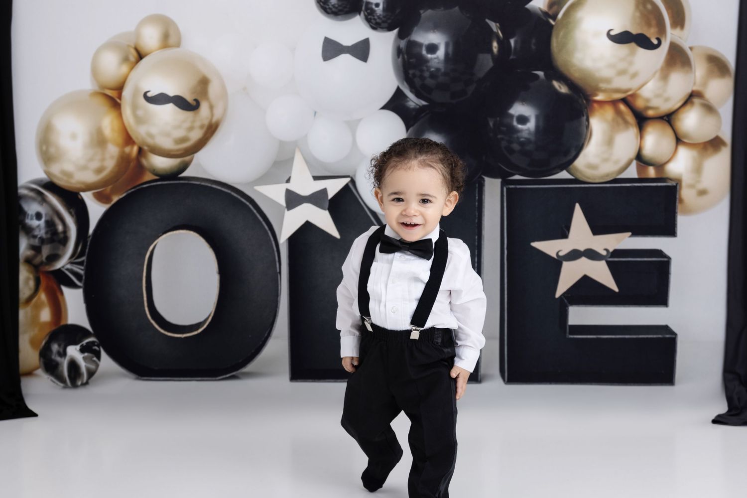 First birthday party setup with black and gold balloon arch, large black ONE letters, and stars against white backdrop.