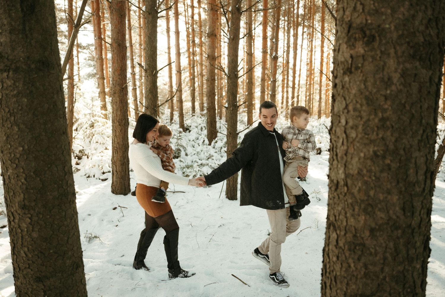 People playfully throwing snow at each other in a sunlit winter forest with tall pine trees.