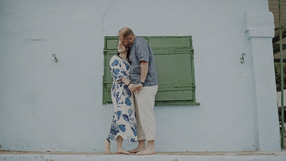 A couple stands against a white wall with green shutters in a Mediterranean setting.