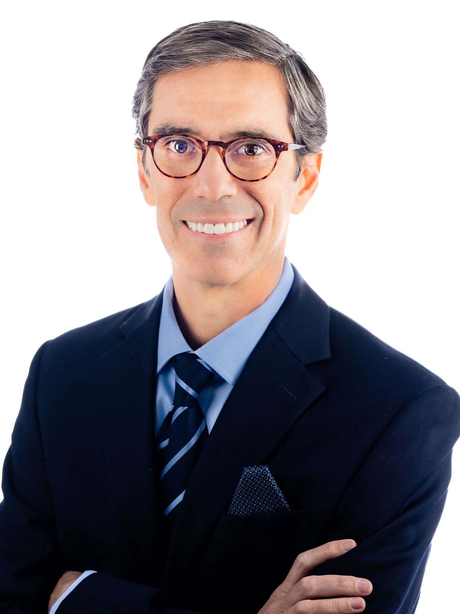 Professional headshot of someone in a navy suit with red-framed glasses, striped tie and pocket square smiling at camera.