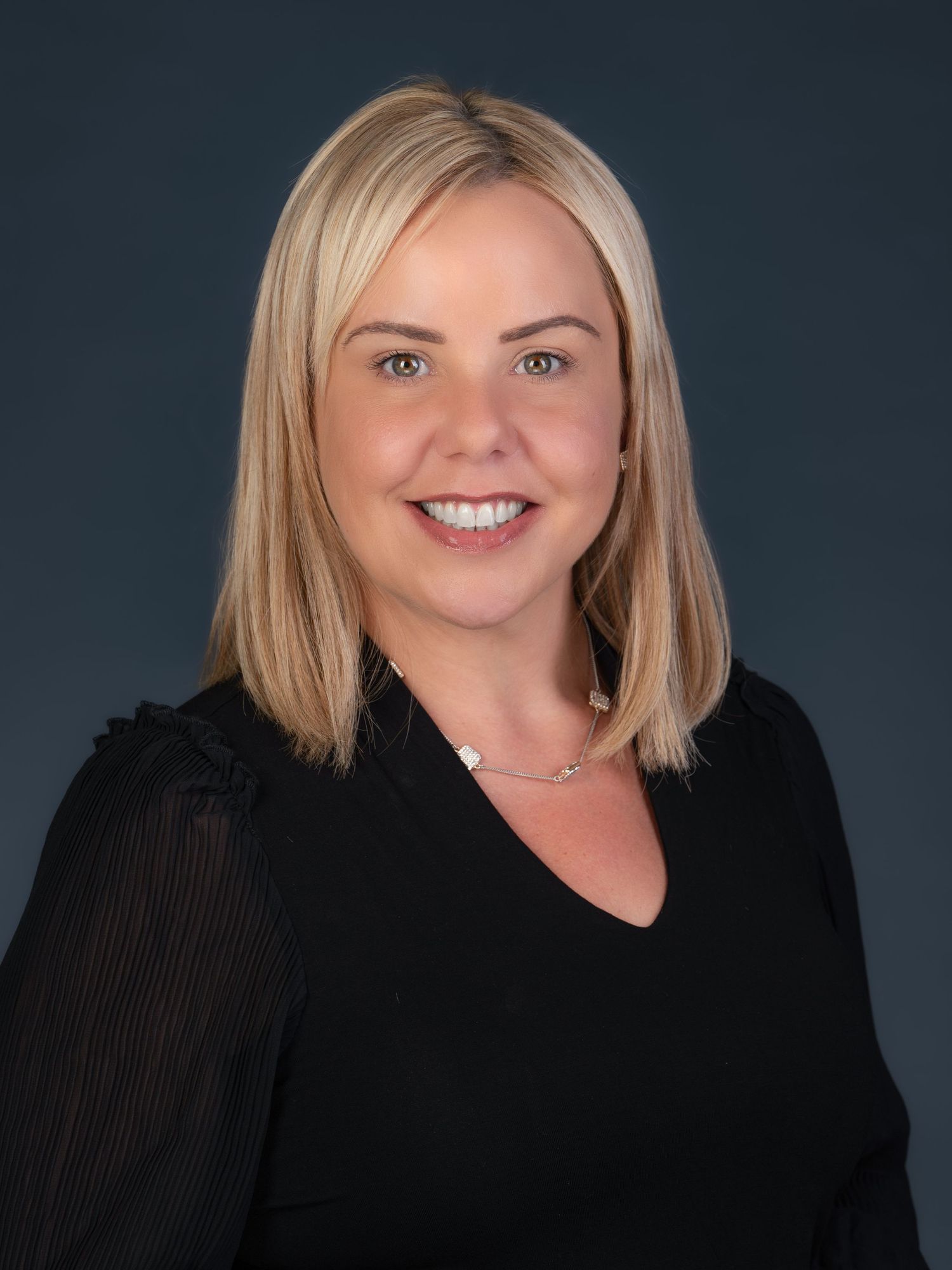 Professional headshot of a person with blonde shoulder-length hair wearing a black v-neck top against a dark background.