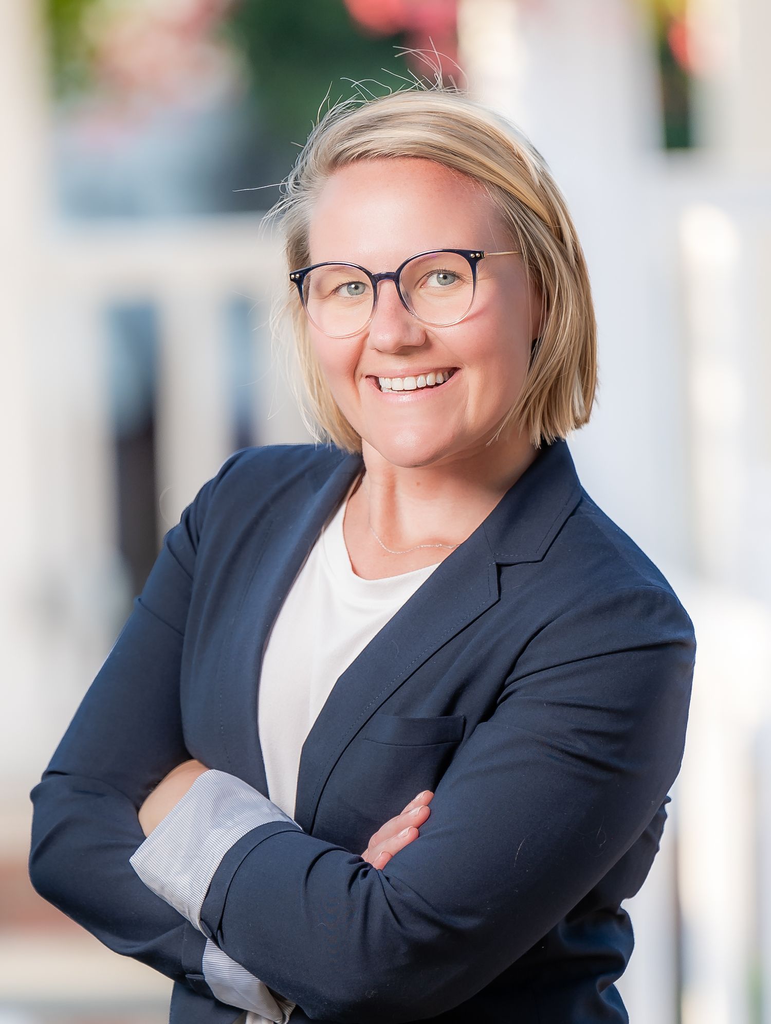 Professional headshot of someone wearing glasses, a navy blazer and white shirt, smiling with arms crossed.