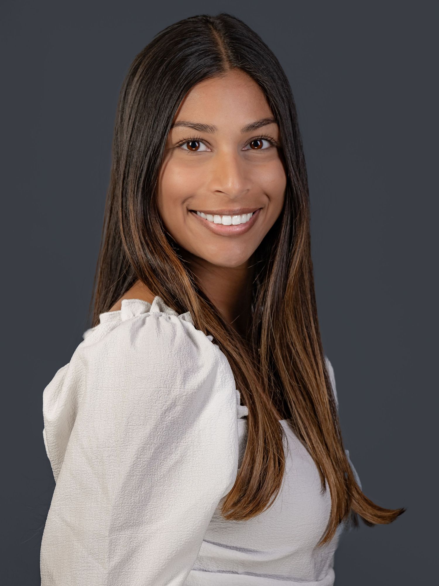 Professional headshot of a smiling person with long brown hair wearing a white top against a dark background.