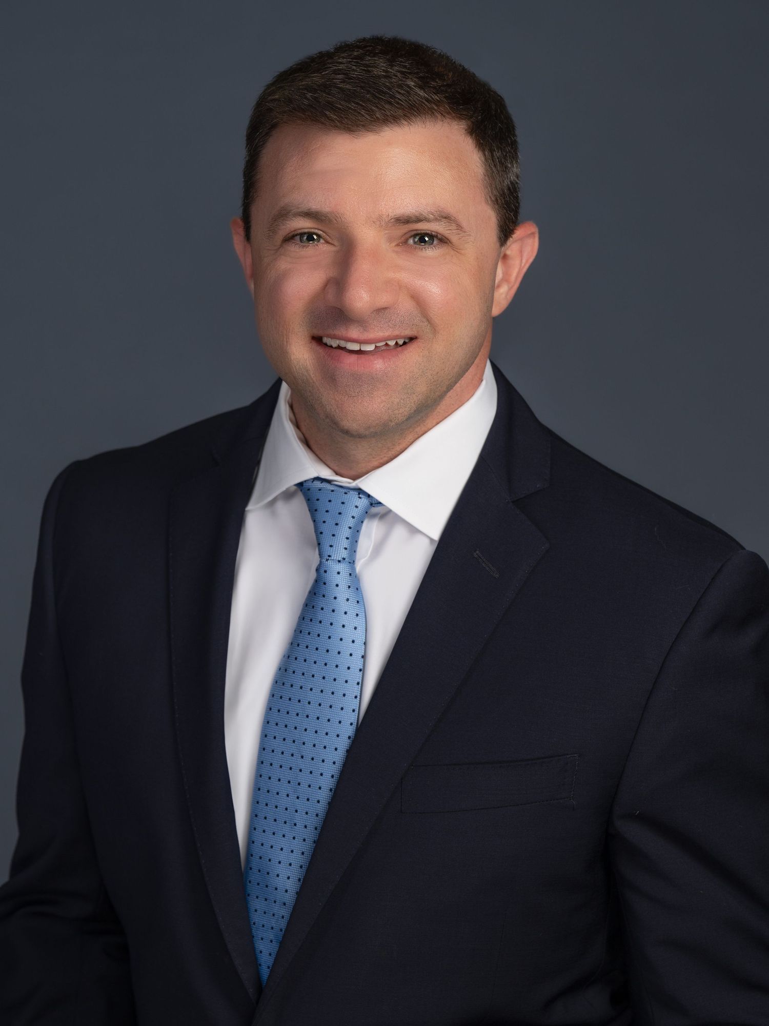 Professional headshot of a person wearing a dark suit and light blue patterned tie against a dark background.