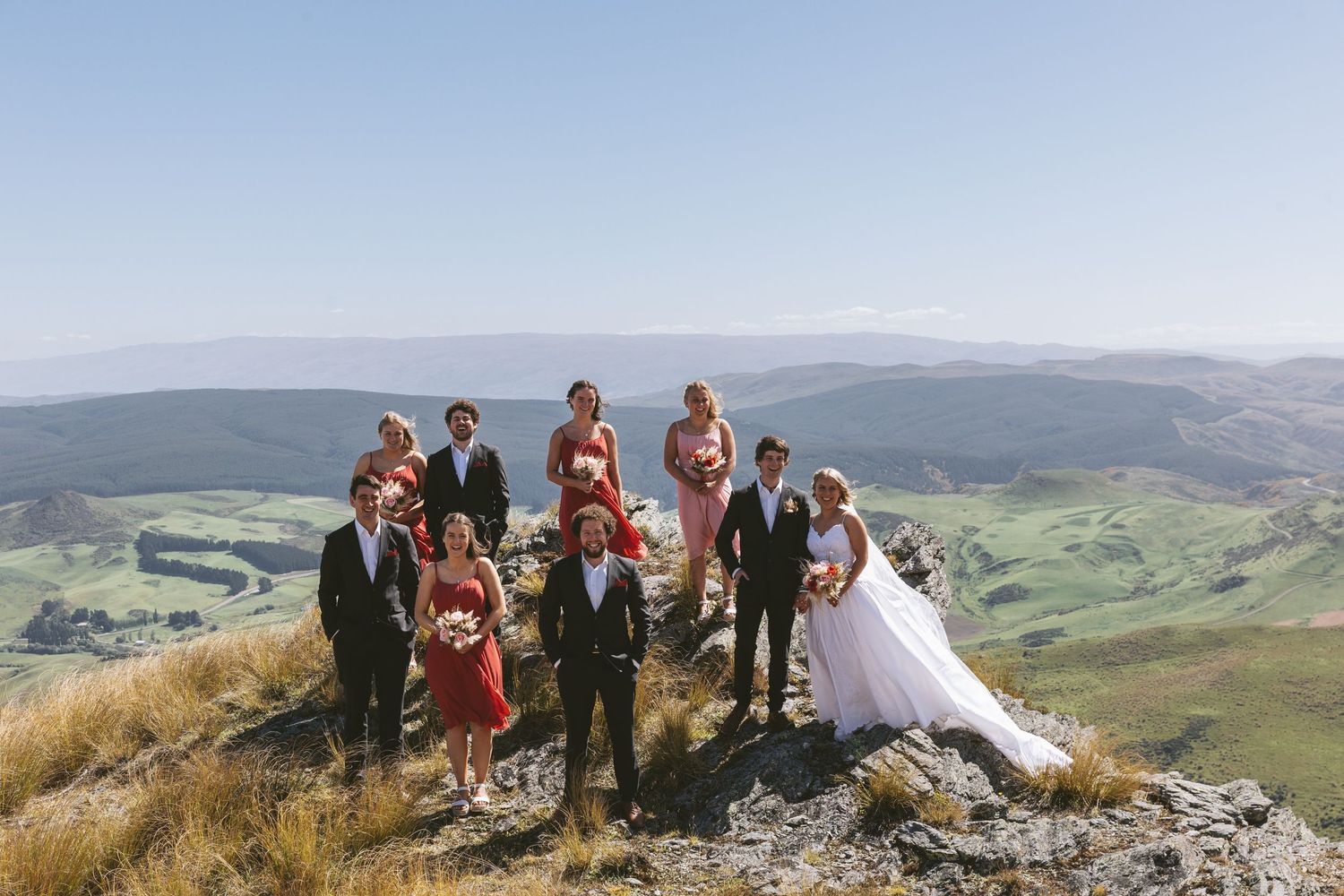 Wedding party gathered on a rocky mountain peak overlooking a vast valley landscape.