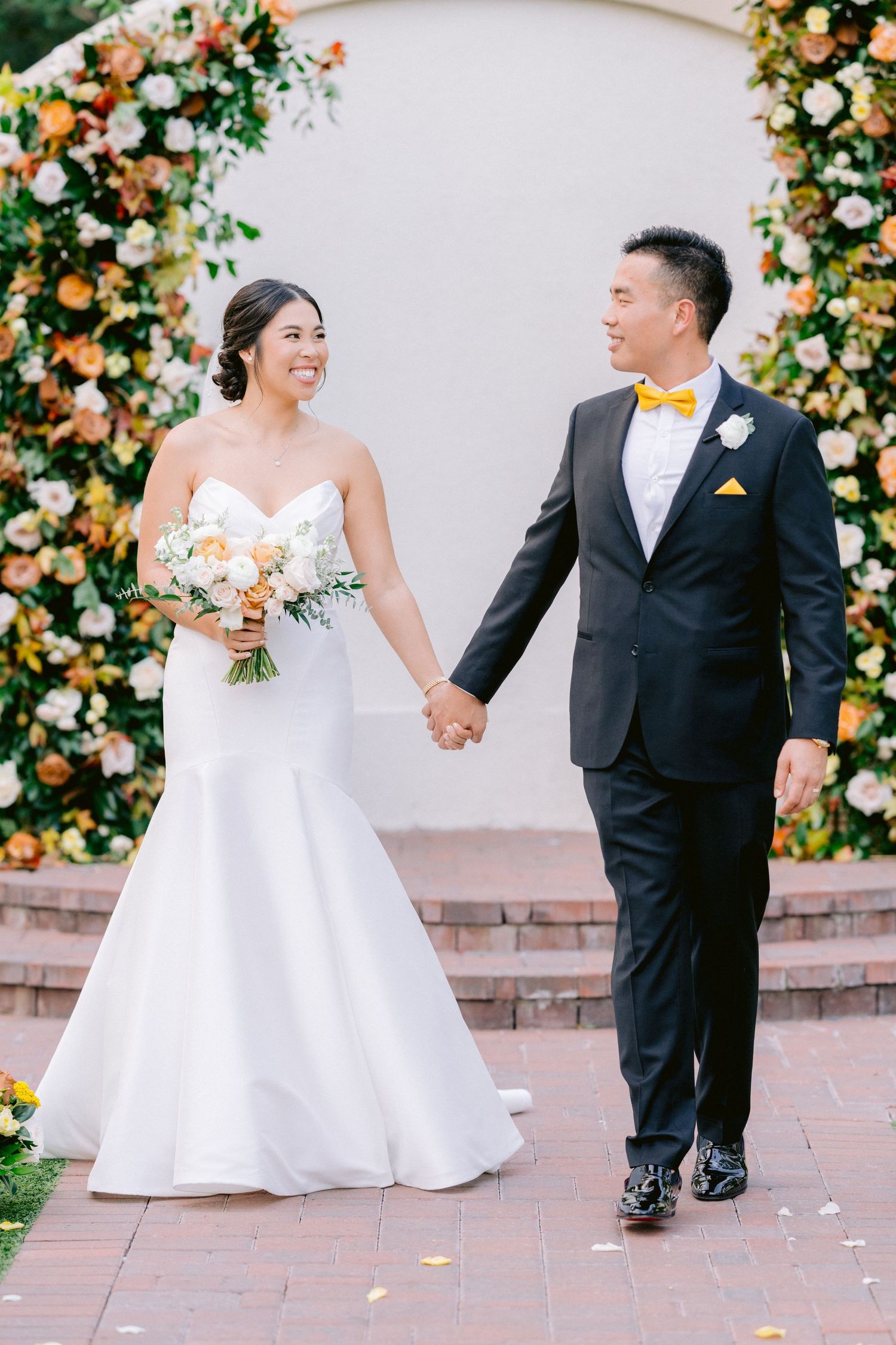 Joyful newlyweds holding hands under floral arch during wedding ceremony.