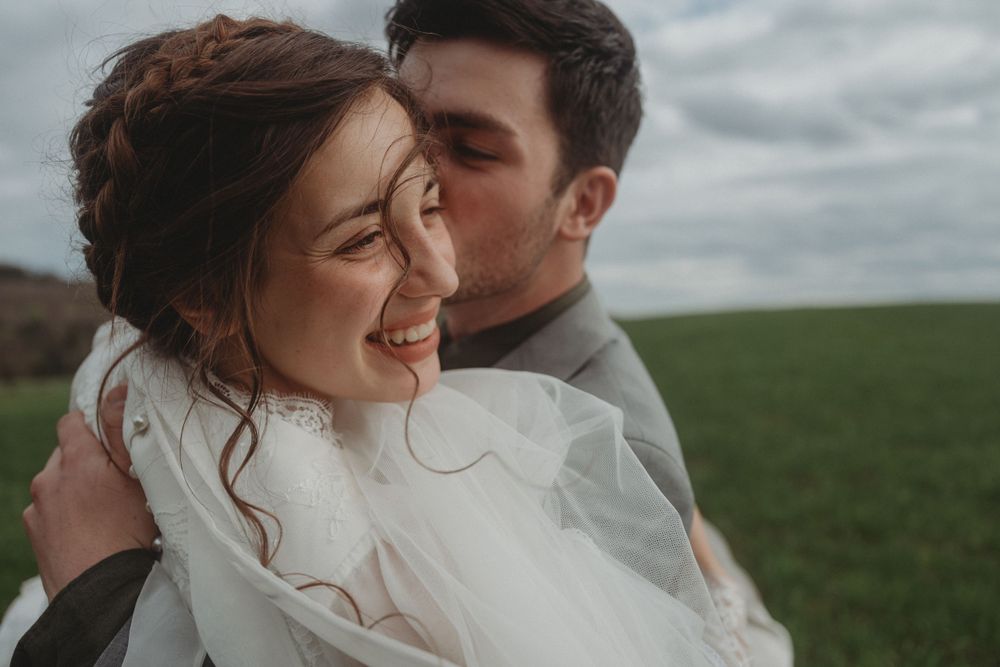 A tender wedding moment captured outdoors in a green field under cloudy skies.