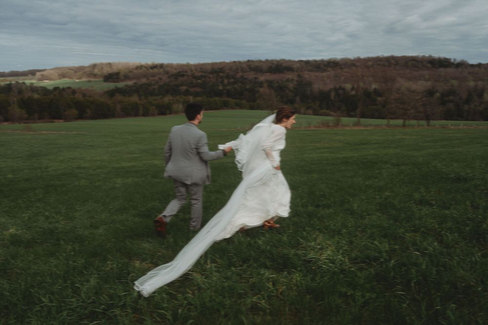 A wedding couple runs playfully through an open field with the bride's dress flowing behind them.