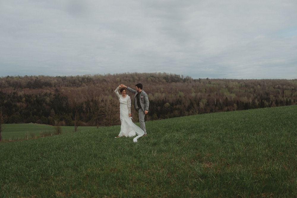 A couple dances joyfully in a green field with mountains in the background on their wedding day.