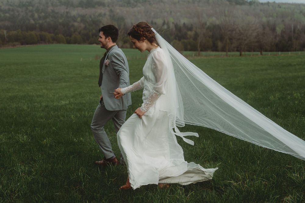 A wedding couple walks through a scenic meadow with the bride's veil flowing in the wind.