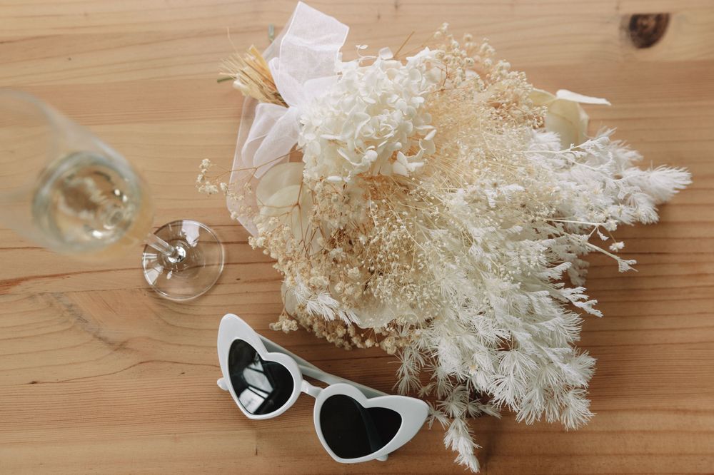 Wedding details with dried flowers, heart sunglasses and champagne glass on wooden surface.