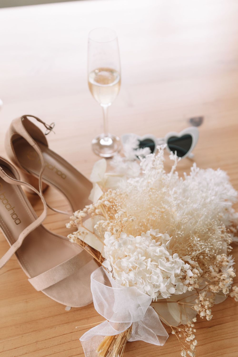 Bridal accessories including nude heels, dried flowers and champagne glass on wooden floor.