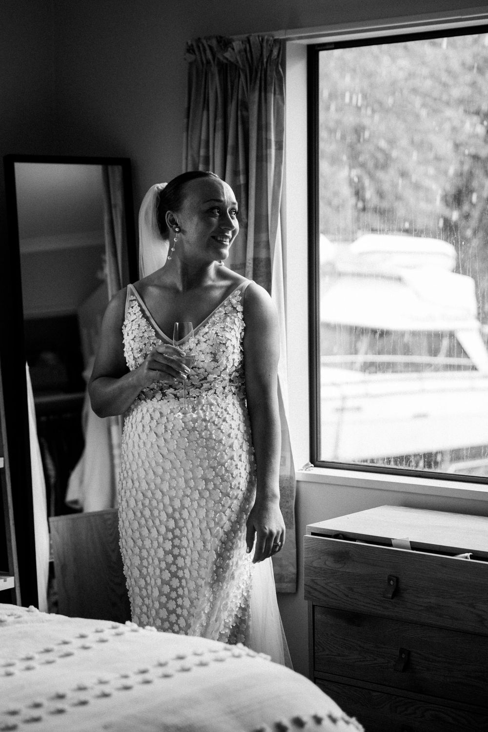 A black and white photograph captures a serene moment by a window in a wedding dress.