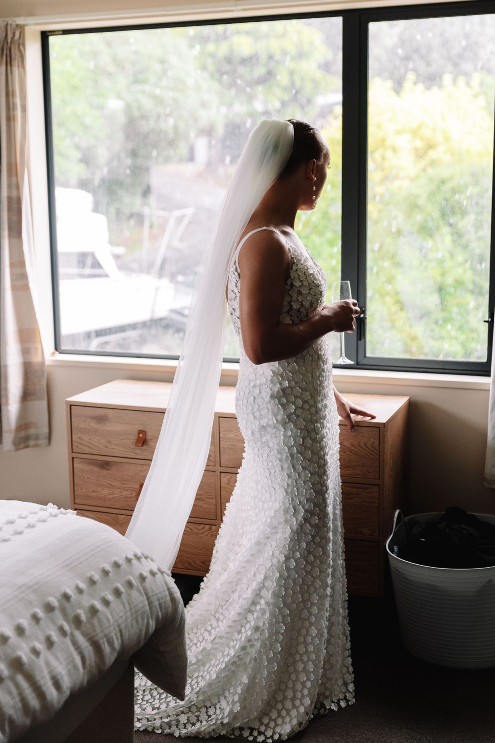A bride in a fitted lace gown gazes out a window, her long veil flowing behind her.
