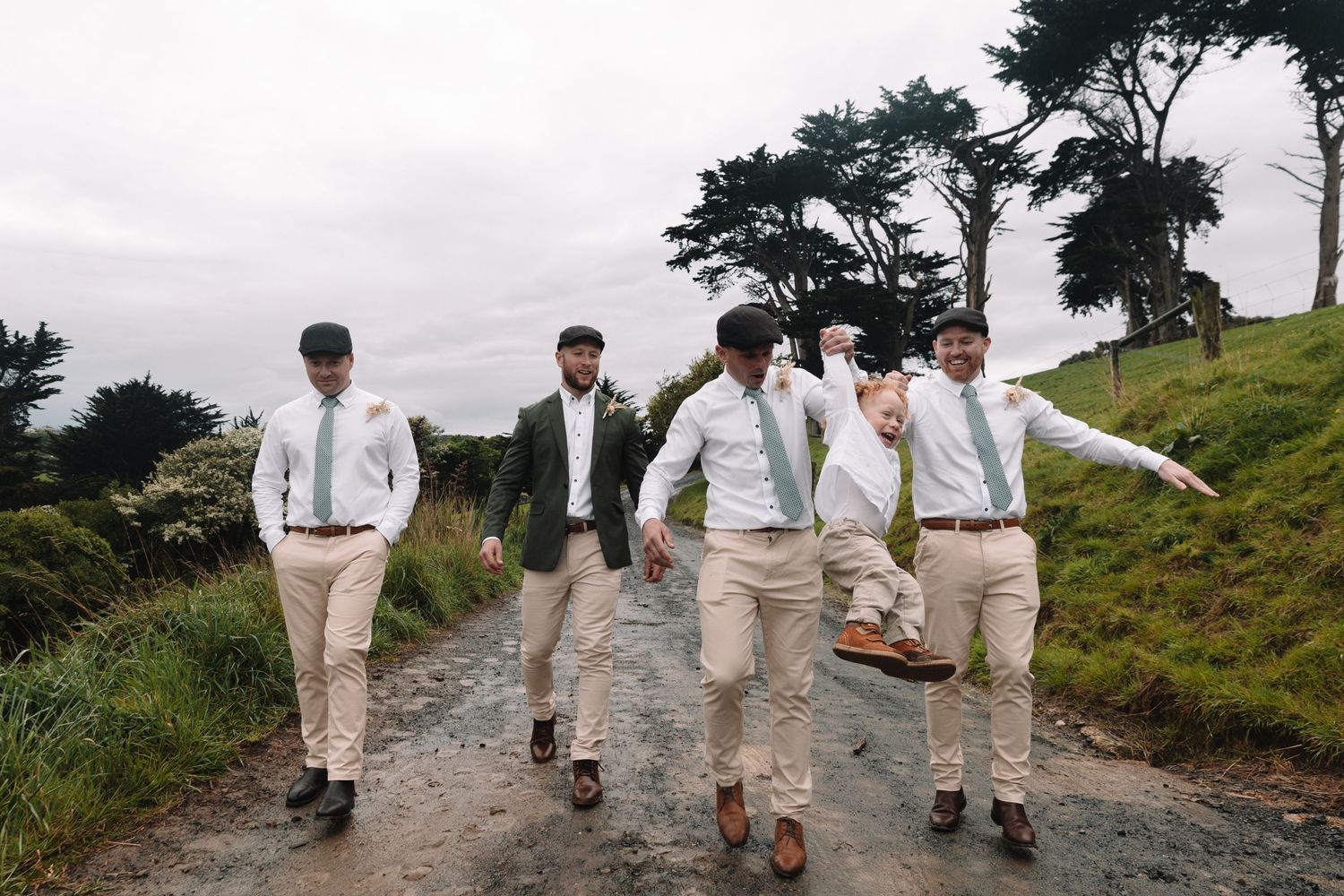 Groomsmen in khaki pants and white shirts walk down a scenic rural path surrounded by greenery.