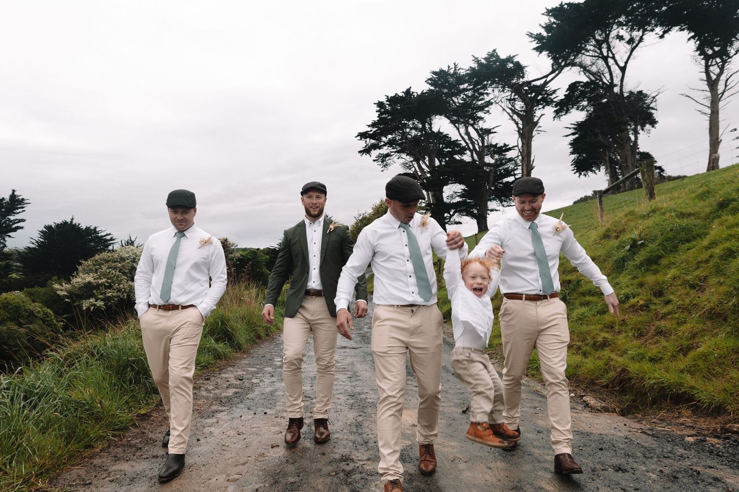 Wedding party members in coordinating outfits jump playfully on a dirt path.