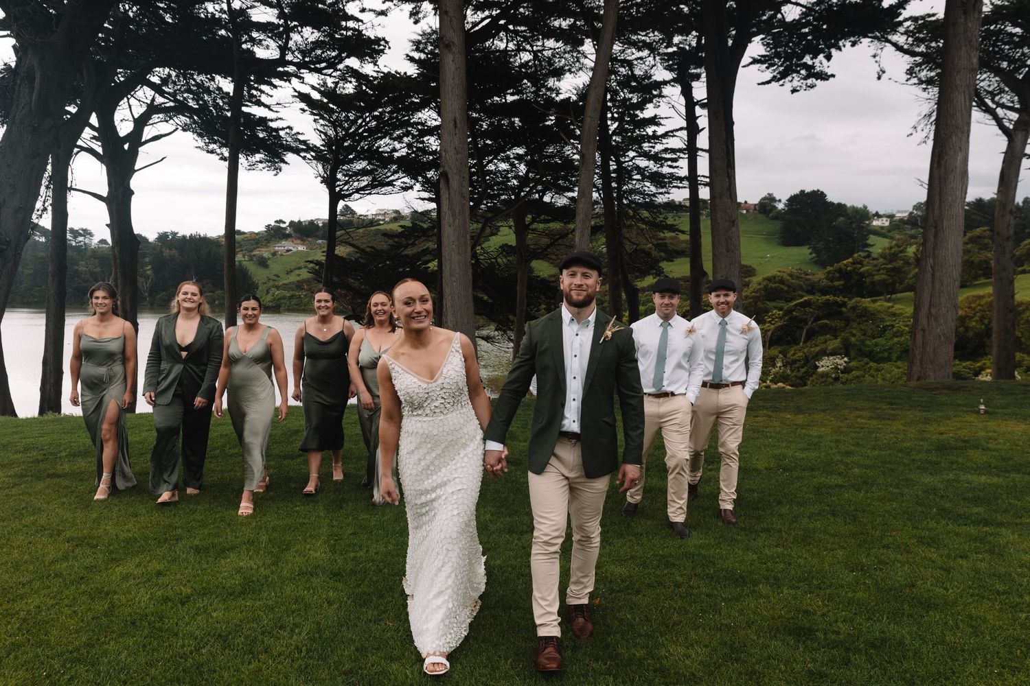 Newlyweds lead their wedding party across a scenic lawn with tall trees and water view.