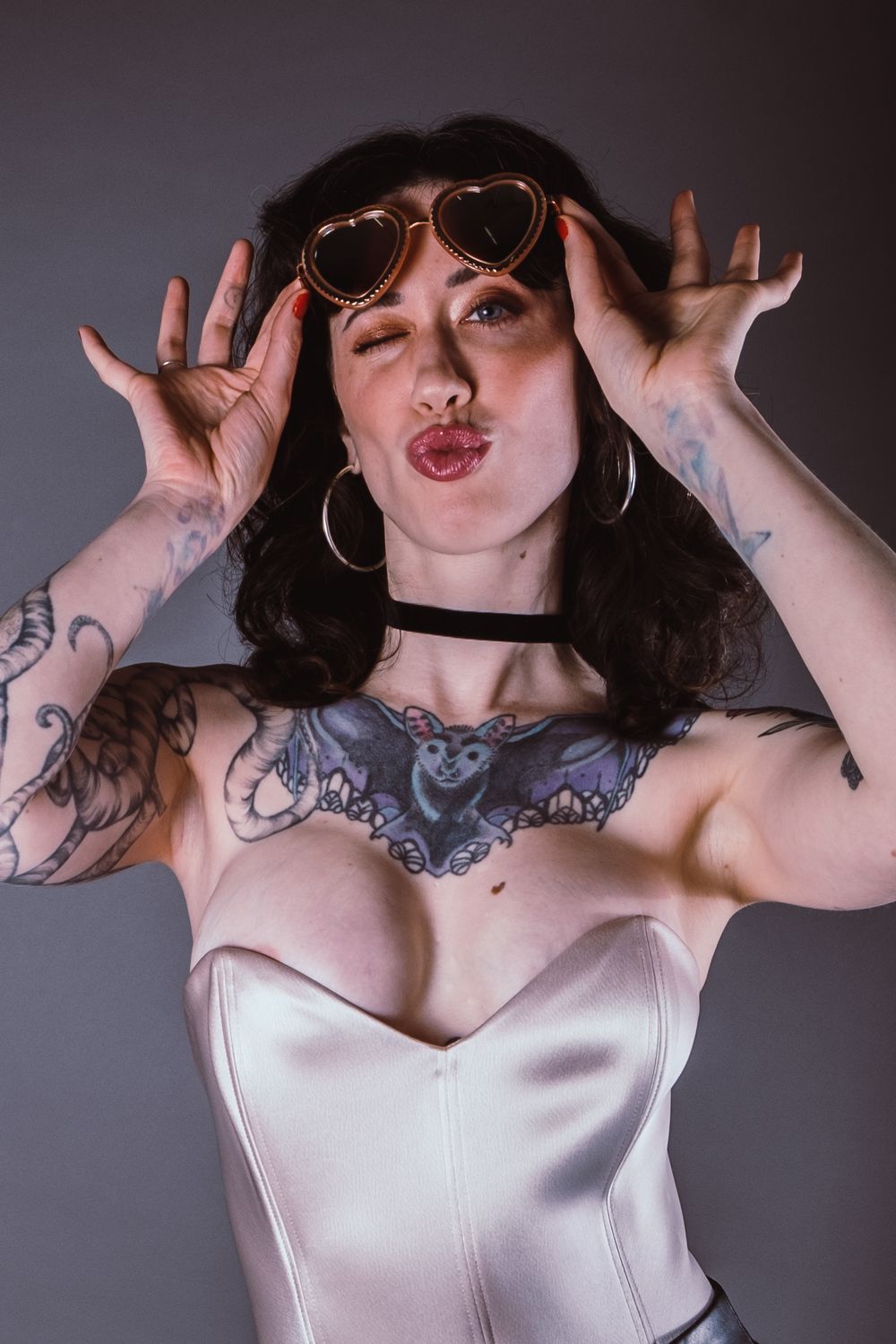 Playful fashion portrait featuring heart sunglasses, tattoos, and a white corset top with expressive pose.