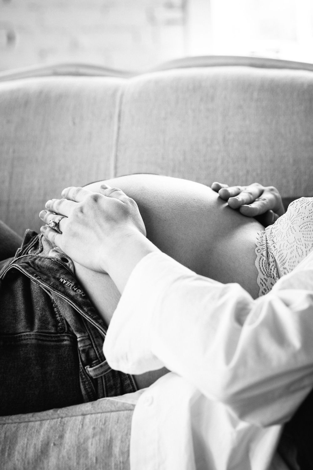 Black and white photo of hands resting on pregnant belly on grey couch.