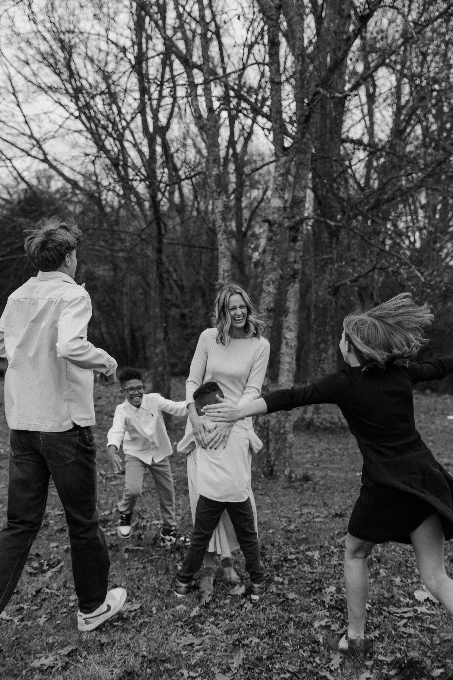 A joyful black and white photograph showing 4 children running to hug their mom outdoors among bare trees in casual attire.