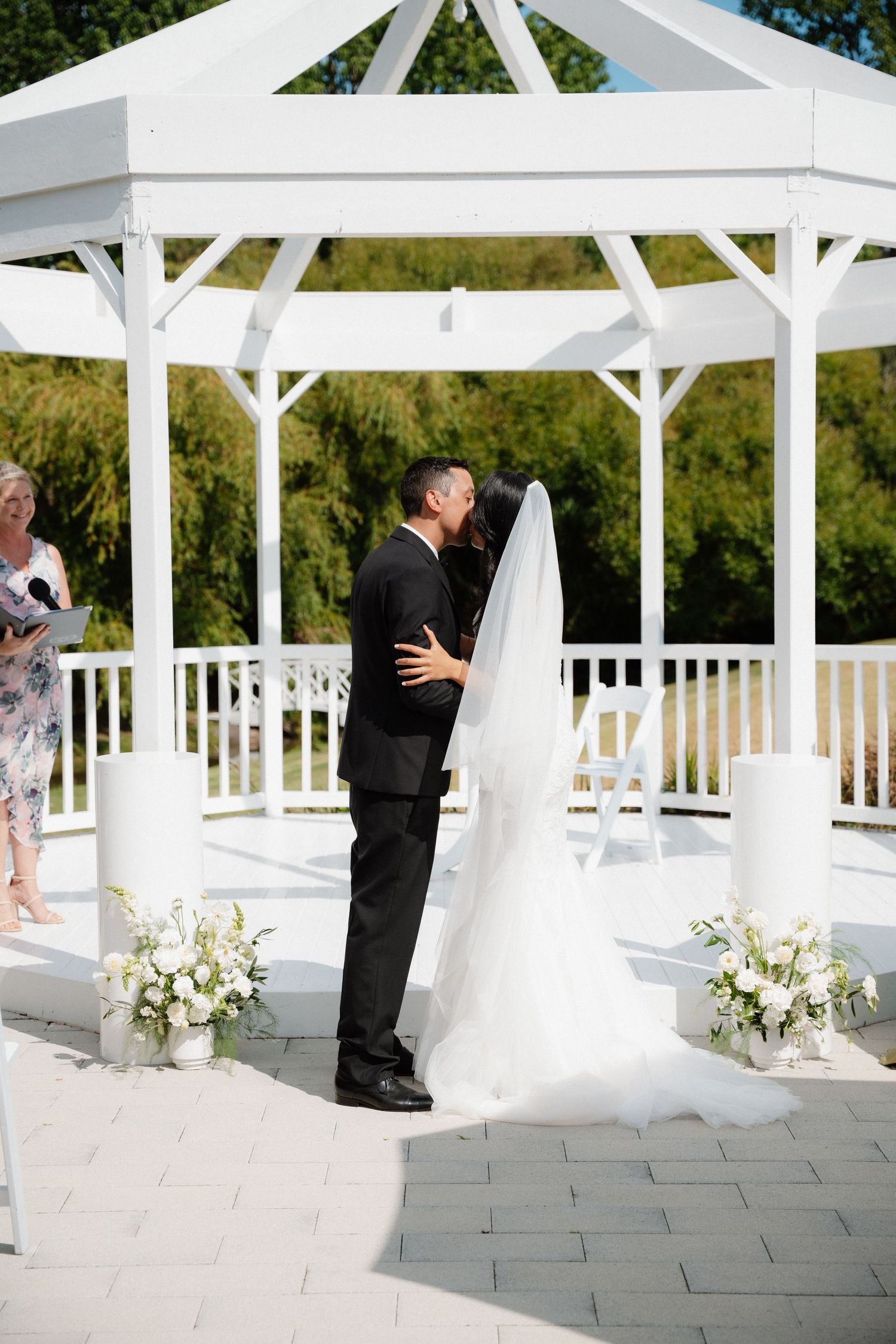 A couple share their first kiss in an elegant white gazebo decorated with floral arrangements at a summer wedding ceremony.