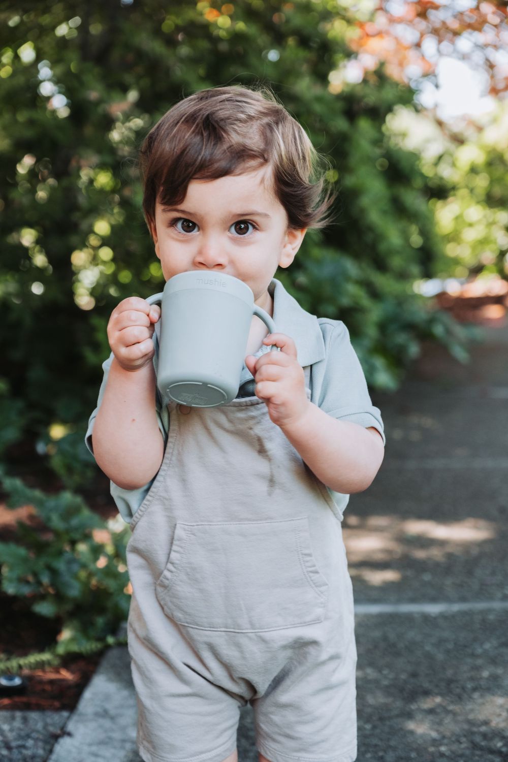 Small child in gray overalls drinks from a light blue mug while standing outdoors on a sunny day.