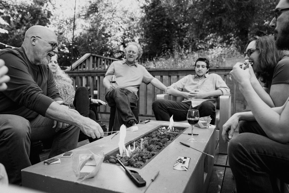 A group of people sit around a picnic table enjoying drinks and conversation in a black and white photo.