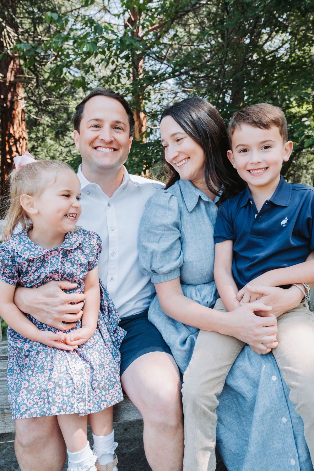 A happy family of four sits together in a casual outdoor setting surrounded by trees and greenery.