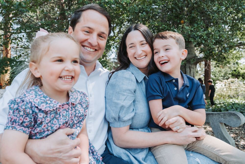 A joyful family of four sits close together outdoors while laughing and smiling in a casual portrait session.