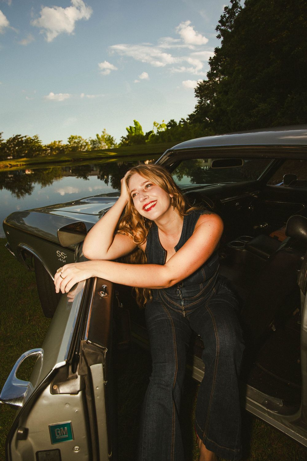 A casual summer scene by a lake with a parked car and someone leaning against its door in golden hour lighting.