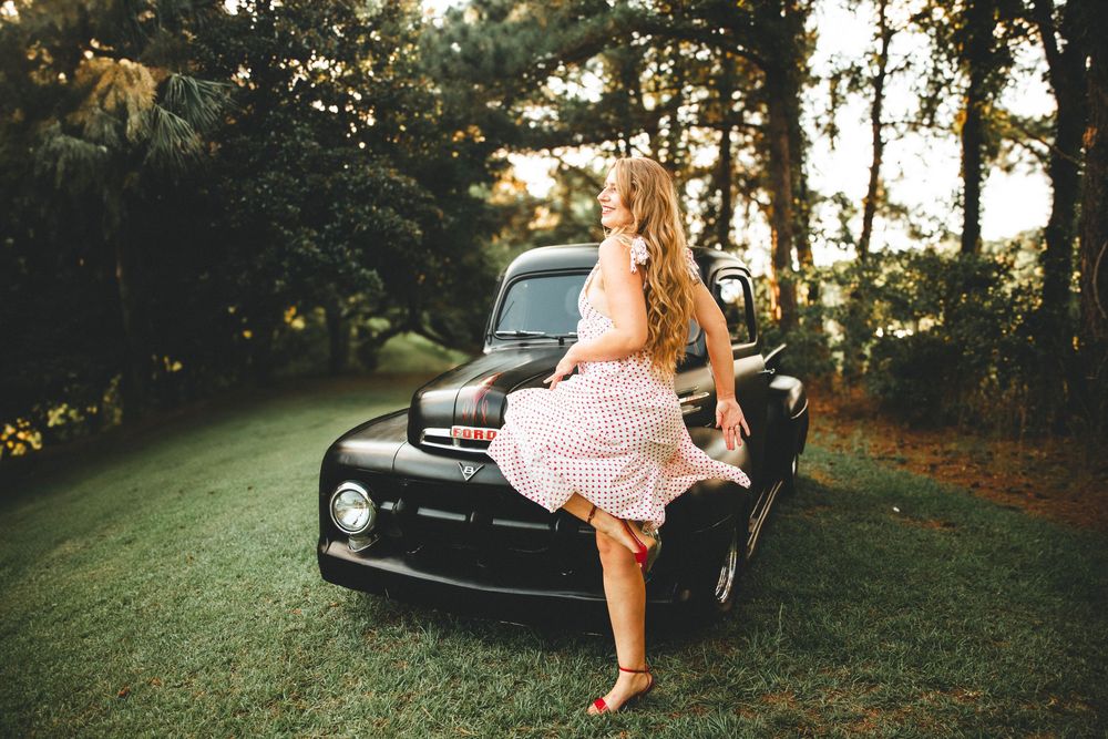 A vintage black Ford truck sits on grass at sunset while someone in a pink polka dot dress leans against the hood.