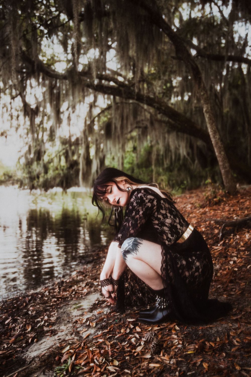 Person in lace dress crouches by a lake shore beneath Spanish moss draped trees in moody atmospheric setting.