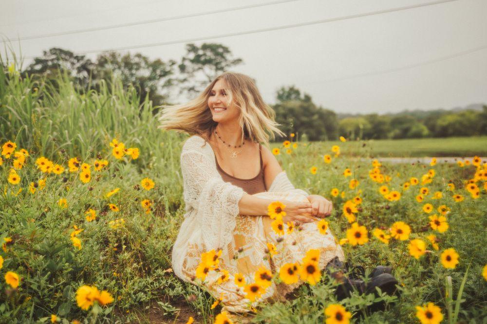 People walk through a vibrant field of yellow sunflowers against a green countryside backdrop on a sunny day.