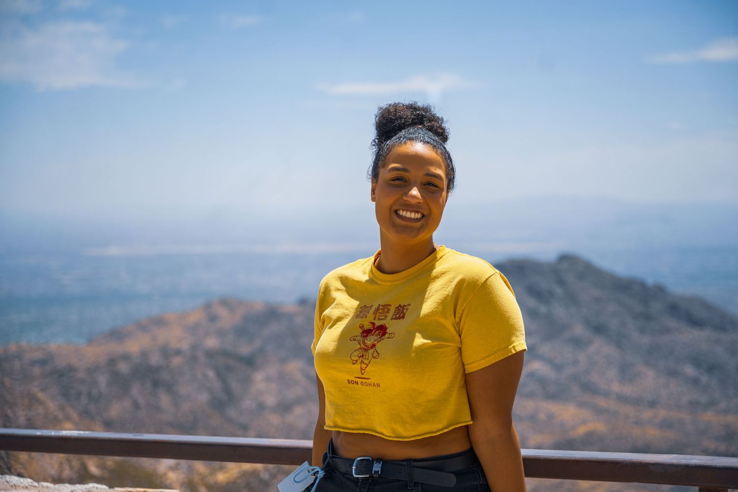 A person in a yellow shirt smiles joyfully while enjoying panoramic mountain views on a clear day.