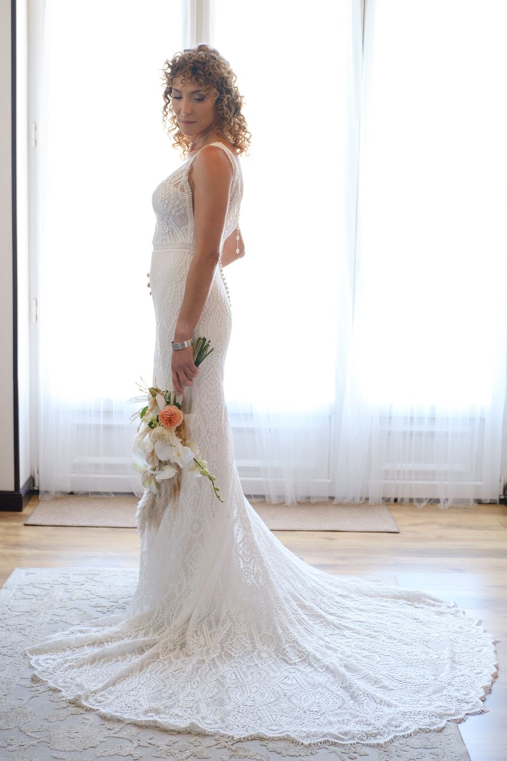 Side profile of a bride in a lace wedding gown holding flowers against a bright window.