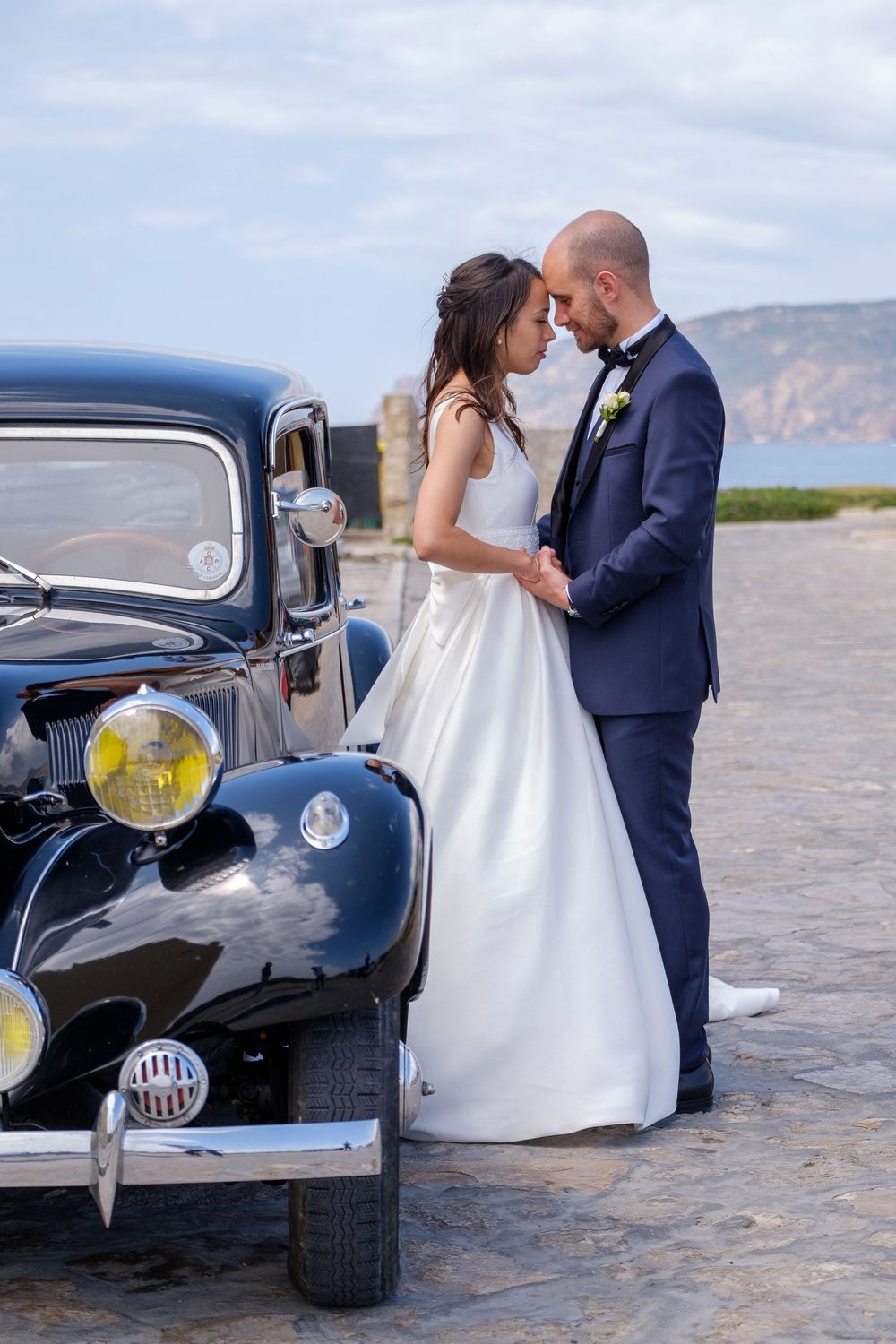 Romantic wedding moments photographed beside a classic black car with ocean views in the background.
