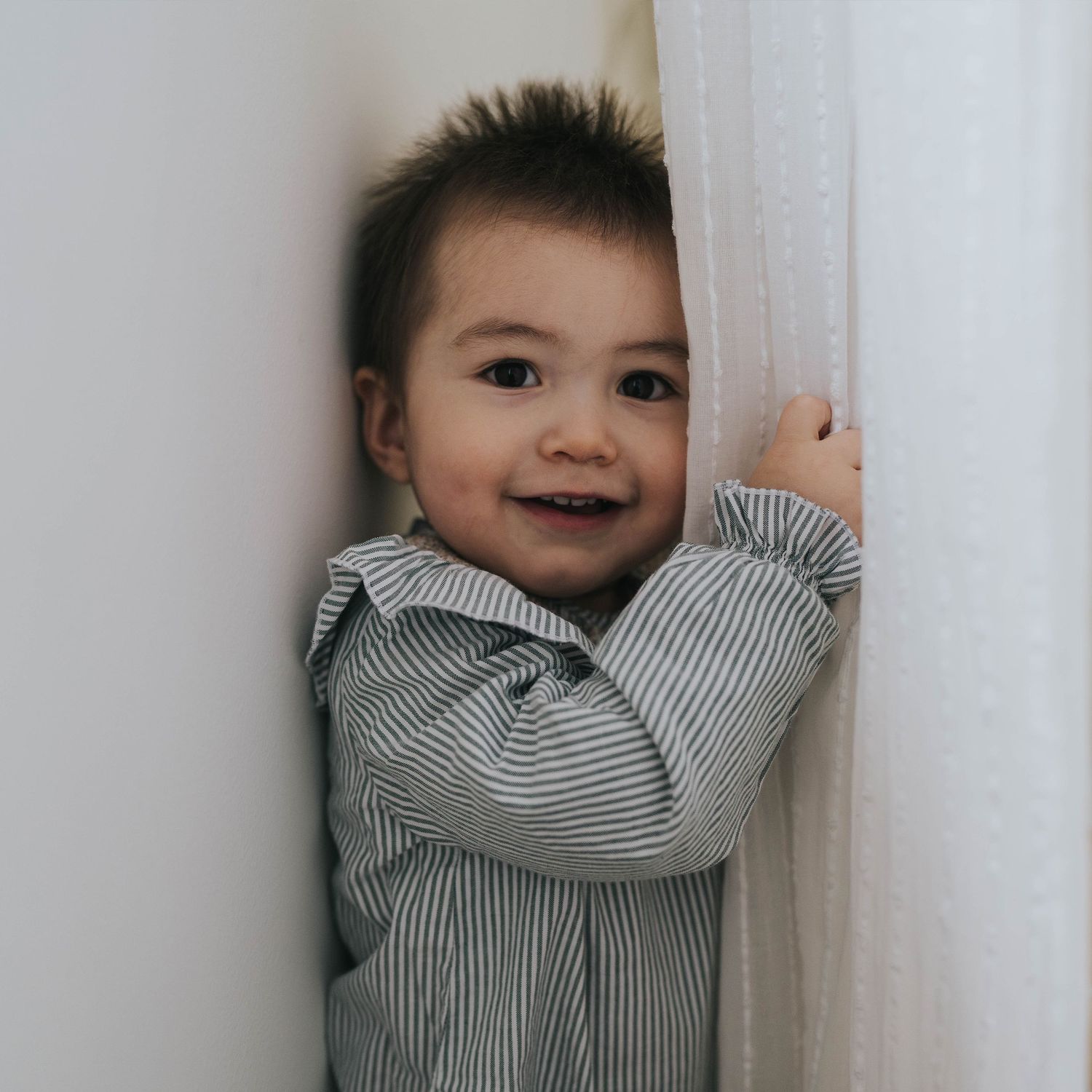 momento cotidiano en su hogar. niño 1 año jugando al escondite con la cortina en su casa en Alcobendas, Madrid, Estaña