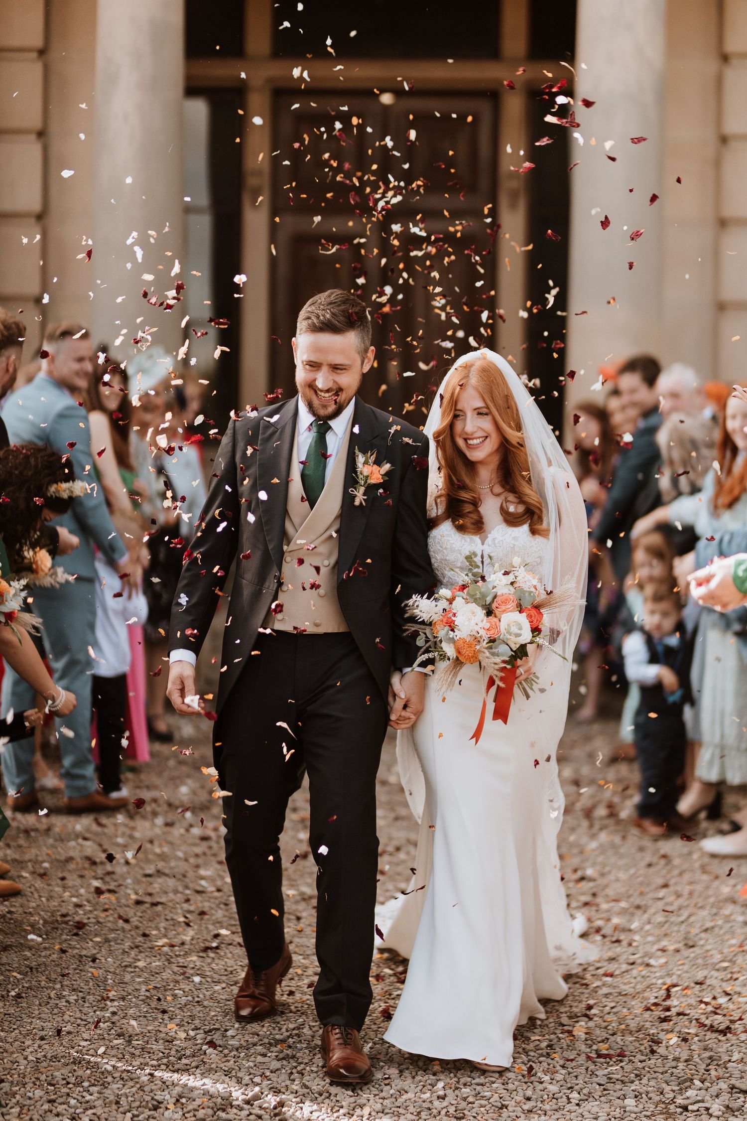 A couple walks through confetti after their wedding ceremony, with her in white dress and him in dark suit with brown shoes.