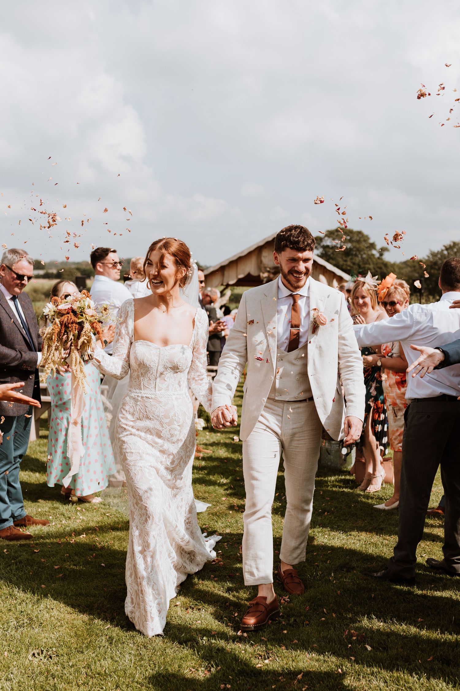 Newlyweds walk through joyful confetti shower on sunny grass lawn as wedding guests celebrate the happy occasion.