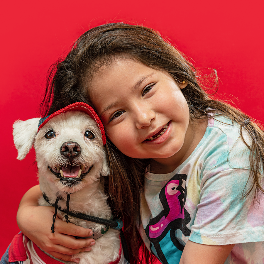 A smiling child hugs their small white dog while posing against a bright red background.