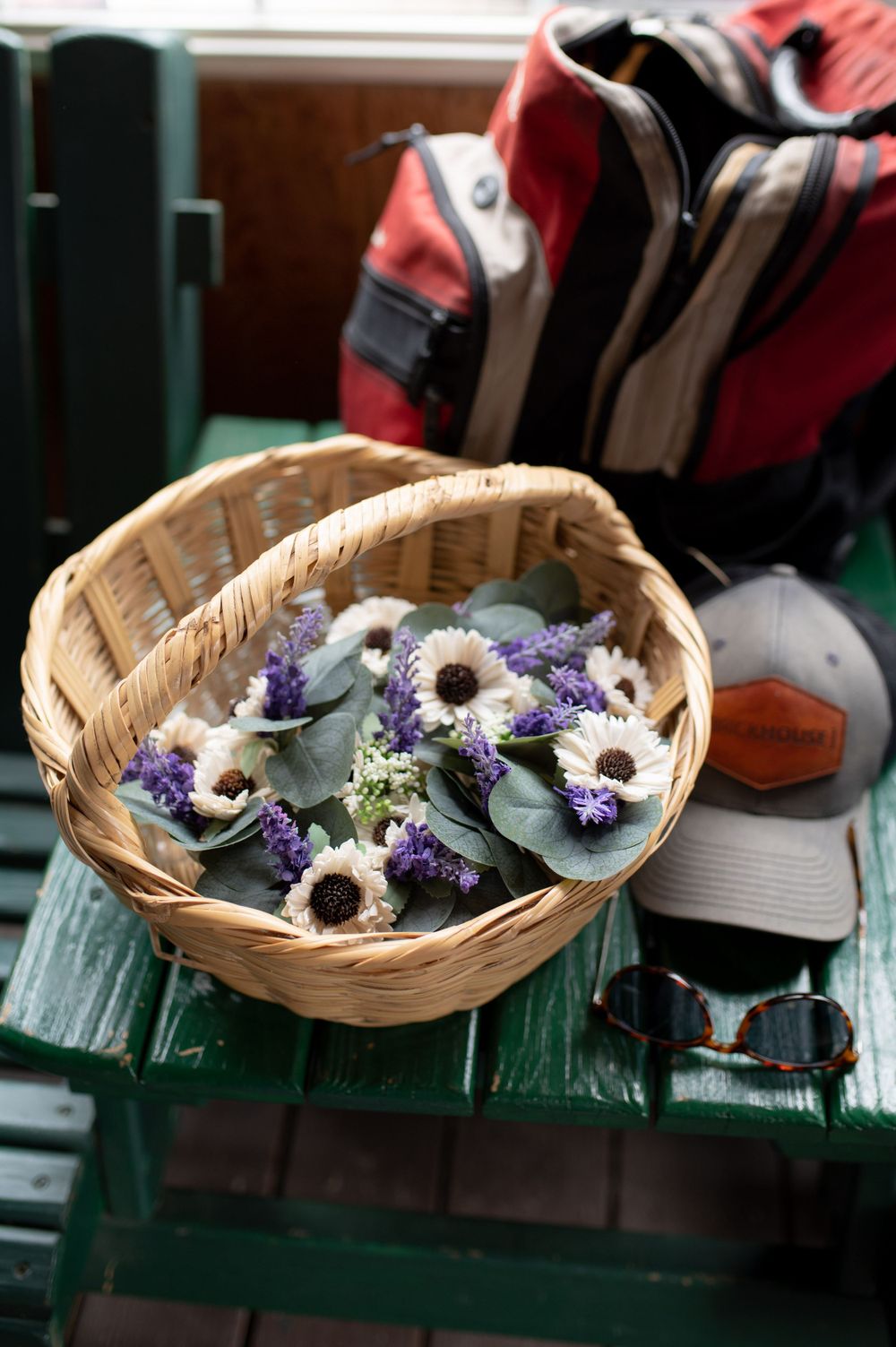 A woven basket filled with purple and white flowers sits near backpacks on a green bench.
