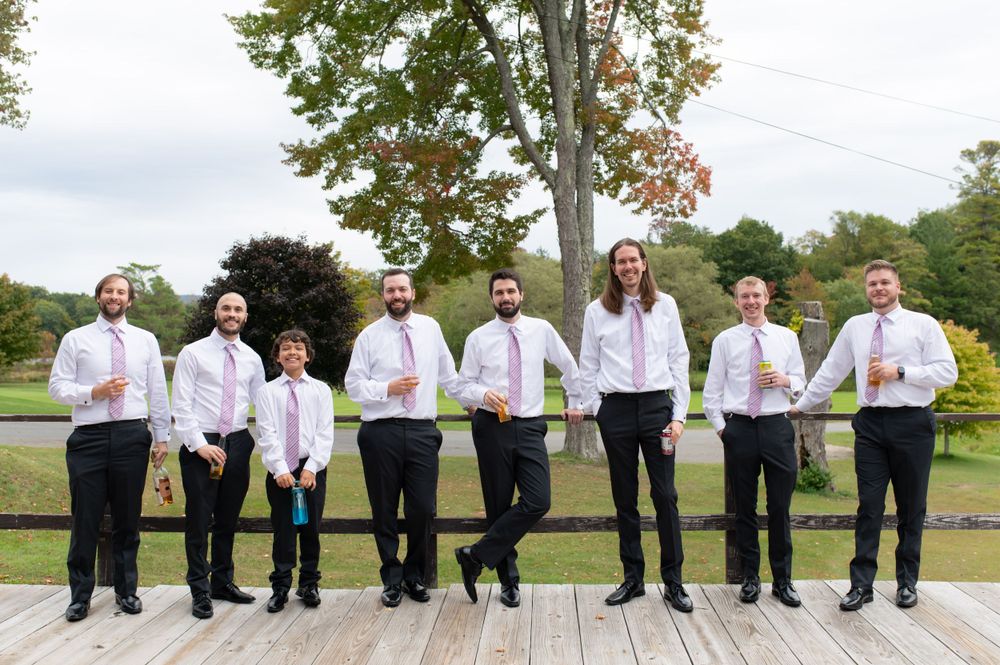 Groomsmen in white shirts and black pants pose together on an outdoor deck for a wedding photo.