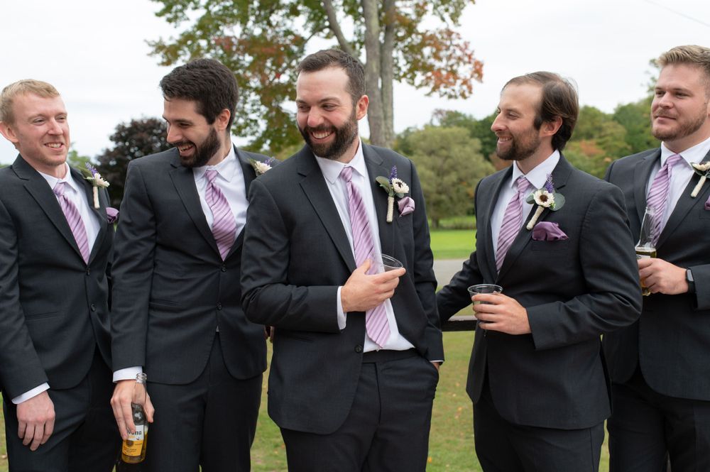 Groomsmen in matching dark suits and pink ties share a laugh outdoors.