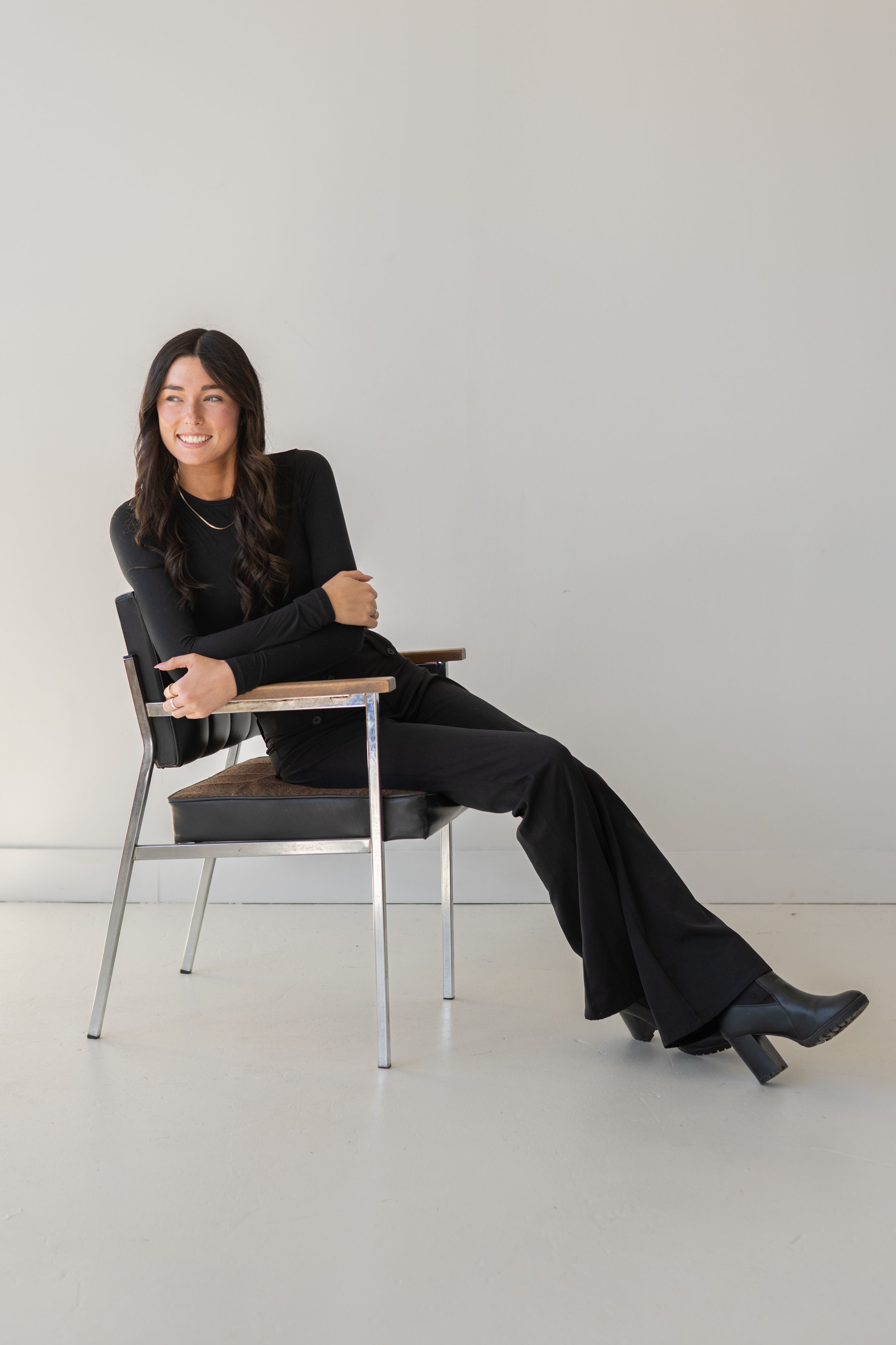 A person in a black outfit sits casually in a modern chair against a light gray wall in a minimalist setting.