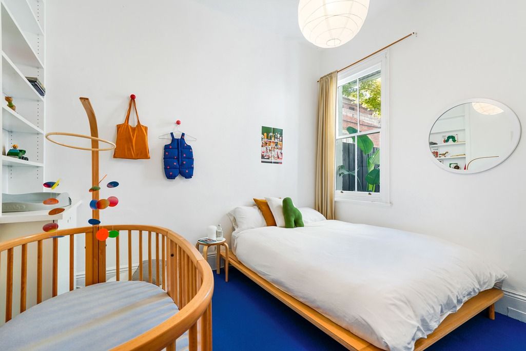 Minimalist bedroom with blue carpet, wooden crib, and white bedding next to large window.