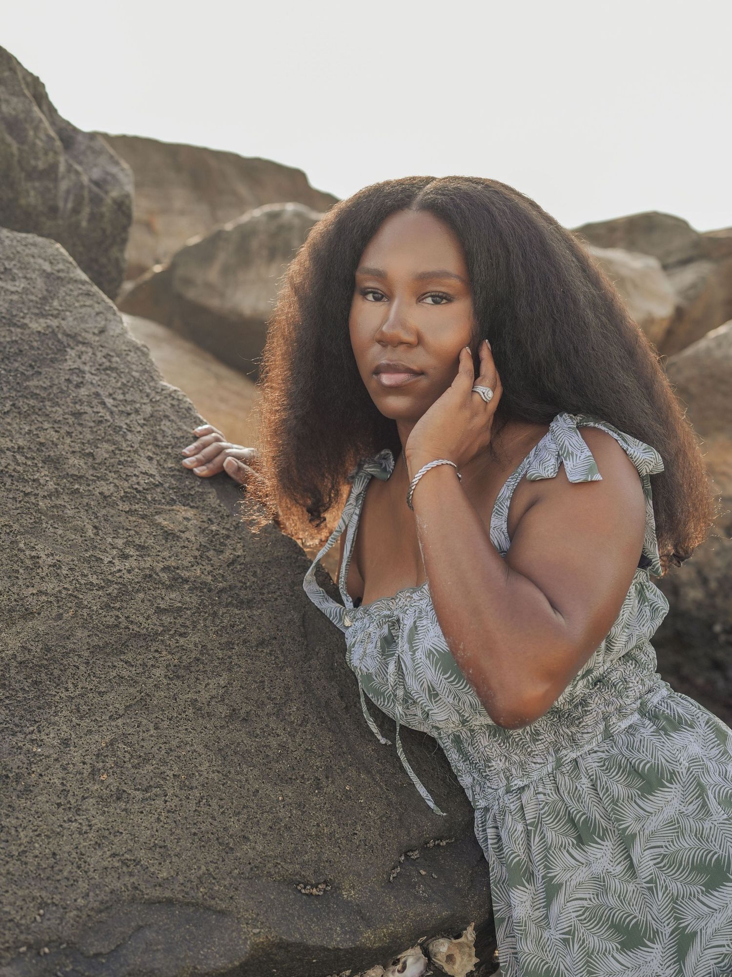A person in a floral dress poses by coastal rocks during golden hour.