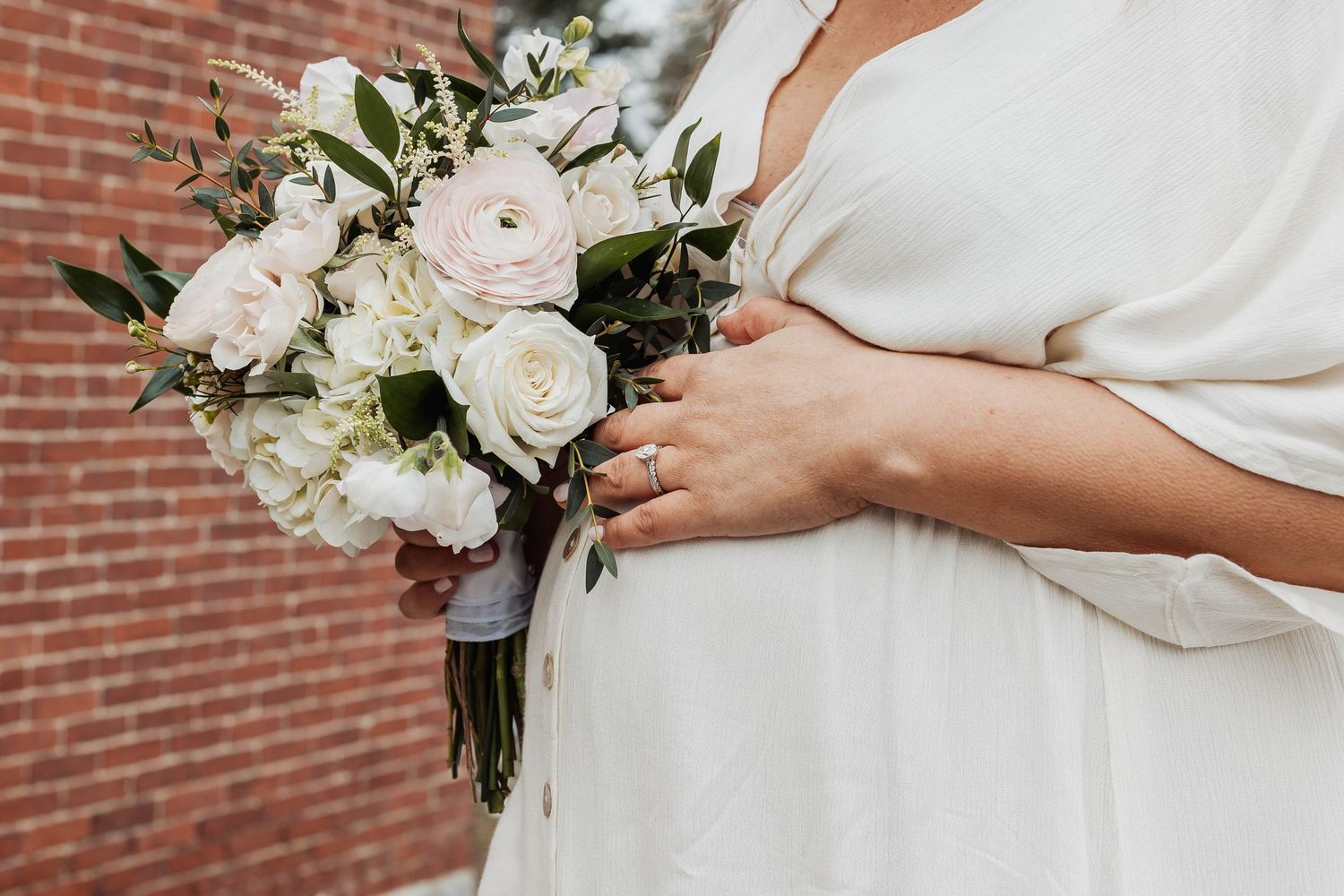 Close-up of a delicate white and blush rose wedding bouquet with greenery held against a white dress.