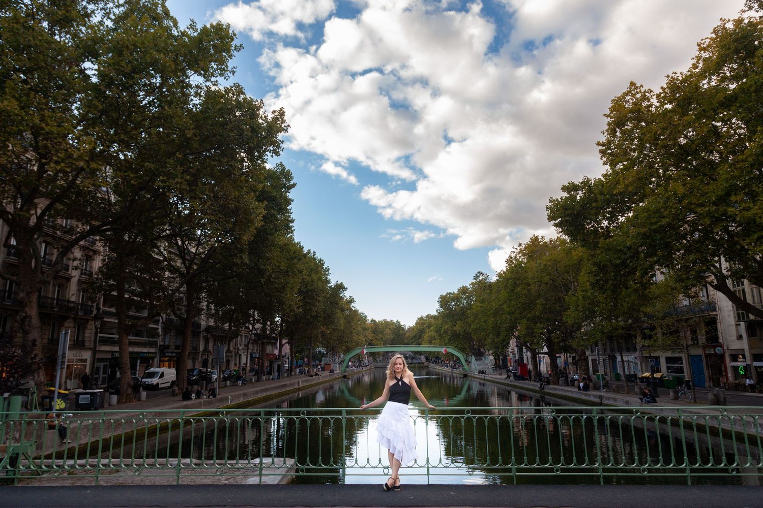 Woman in white dress stands on bridge overlooking scenic canal with trees and buildings in Paris.